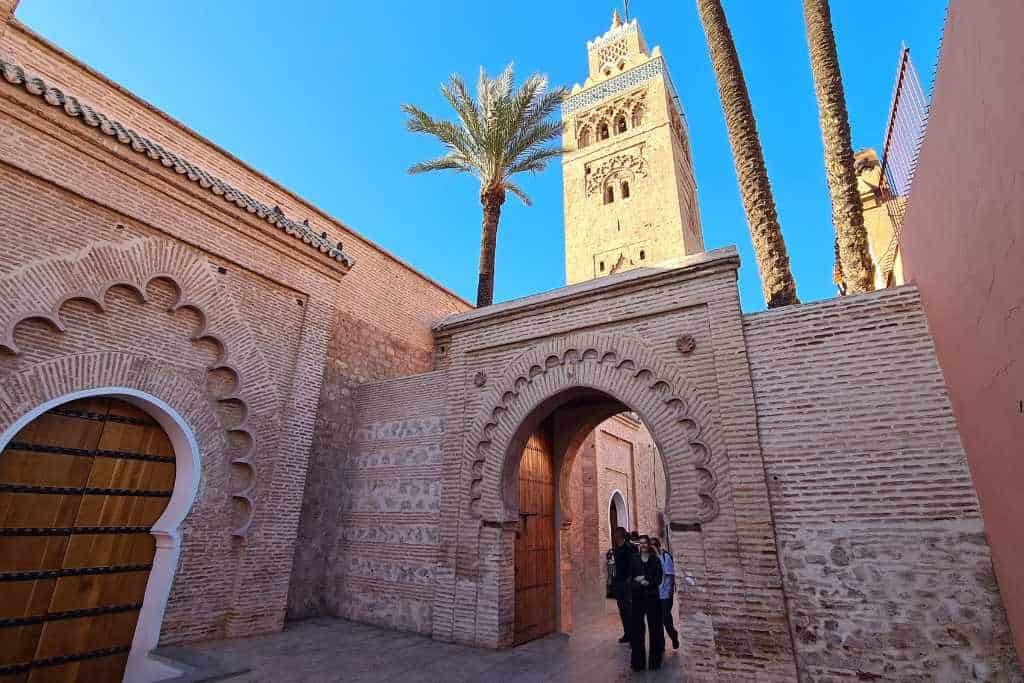 People walking safely under and archway with a tall tower and palm trees in Marrakech.